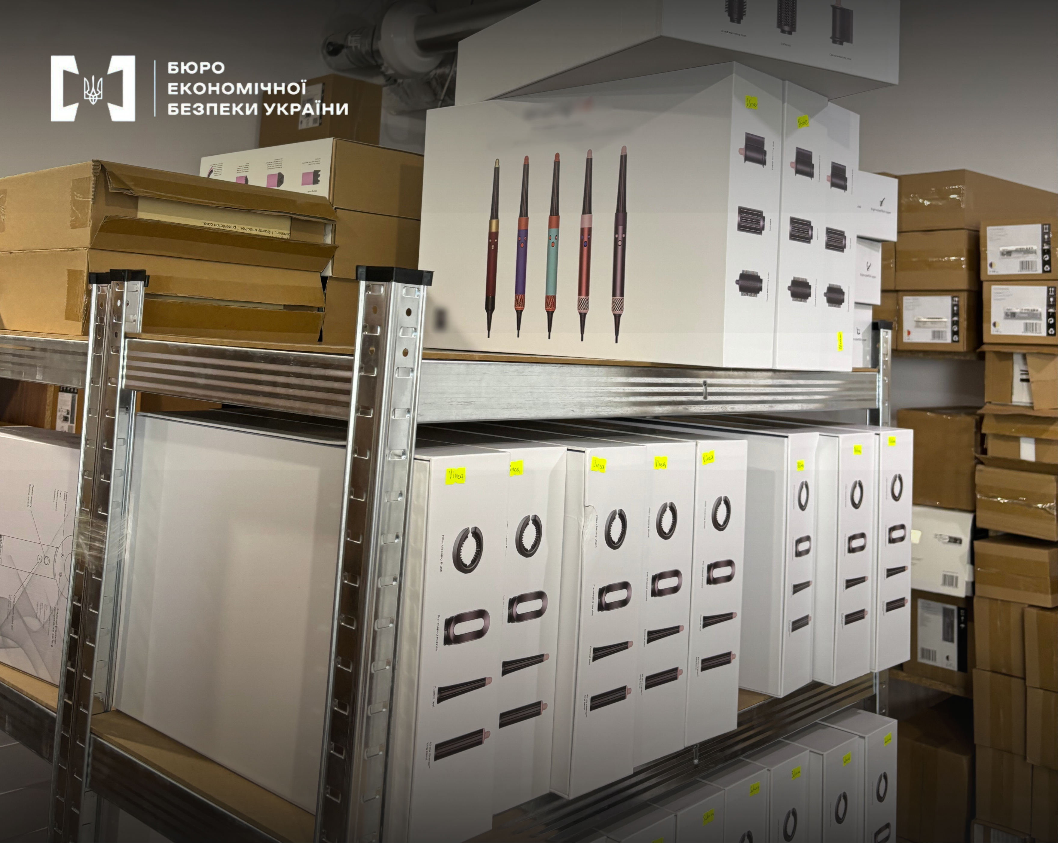 A warehouse with metal shelves filled with numerous cardboard boxes. In the foreground, white boxes with images of stylers in different colors are visible.