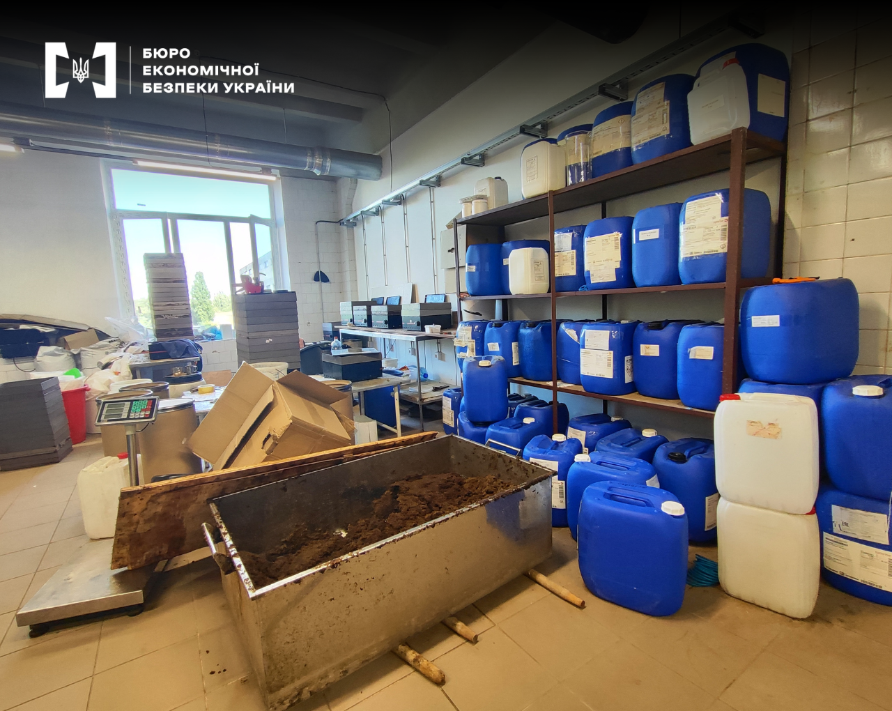 A production facility with numerous plastic canisters, boxes, and a large container with a brown mass.