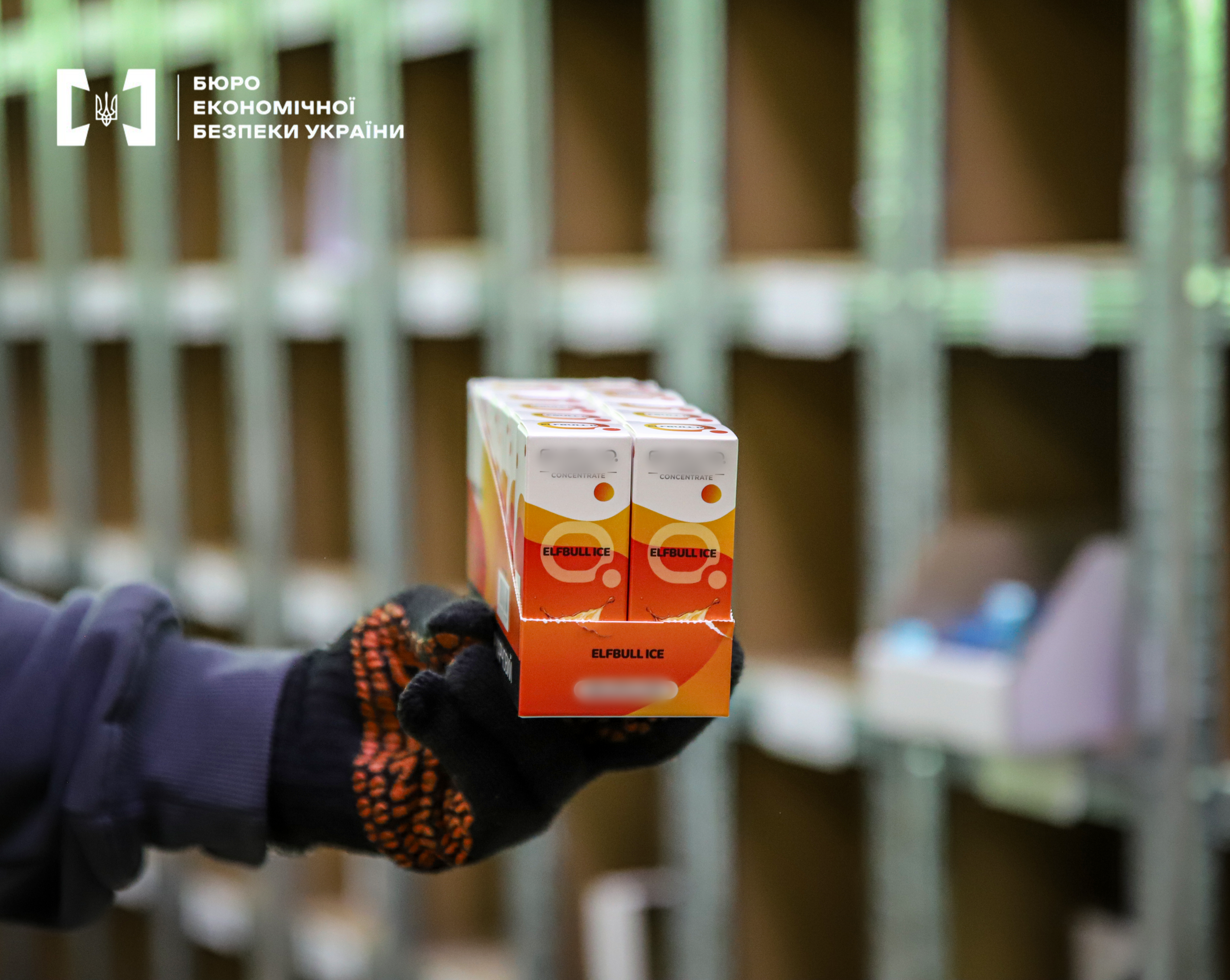 A hand in a protective glove holding a pack of e-liquid for electronic cigarettes. The packaging is a vibrant orange-yellow color with the inscription 'ELFBULL ICE'. The background is blurred