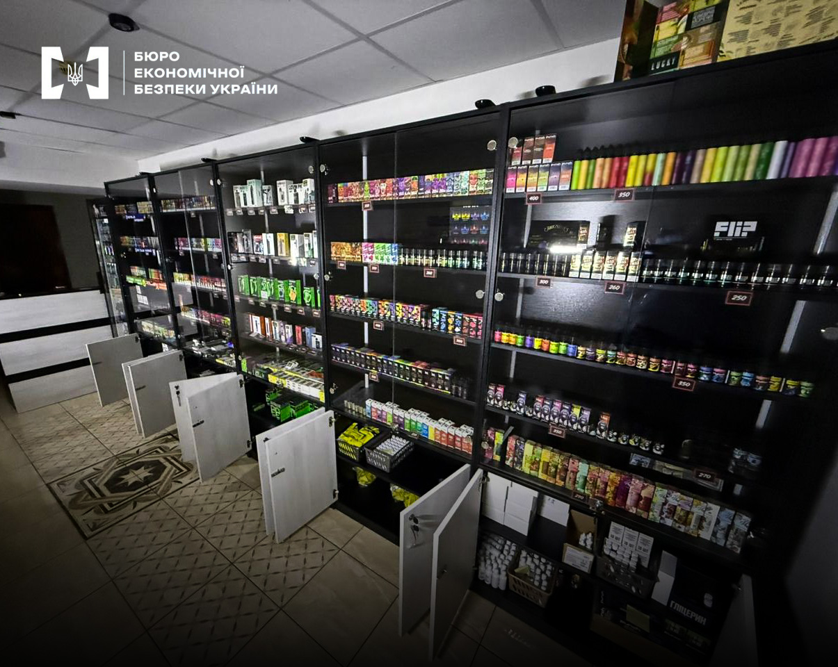 A shop with black shelving units filled with multi-colored bottles and disposable vapes with price tags.