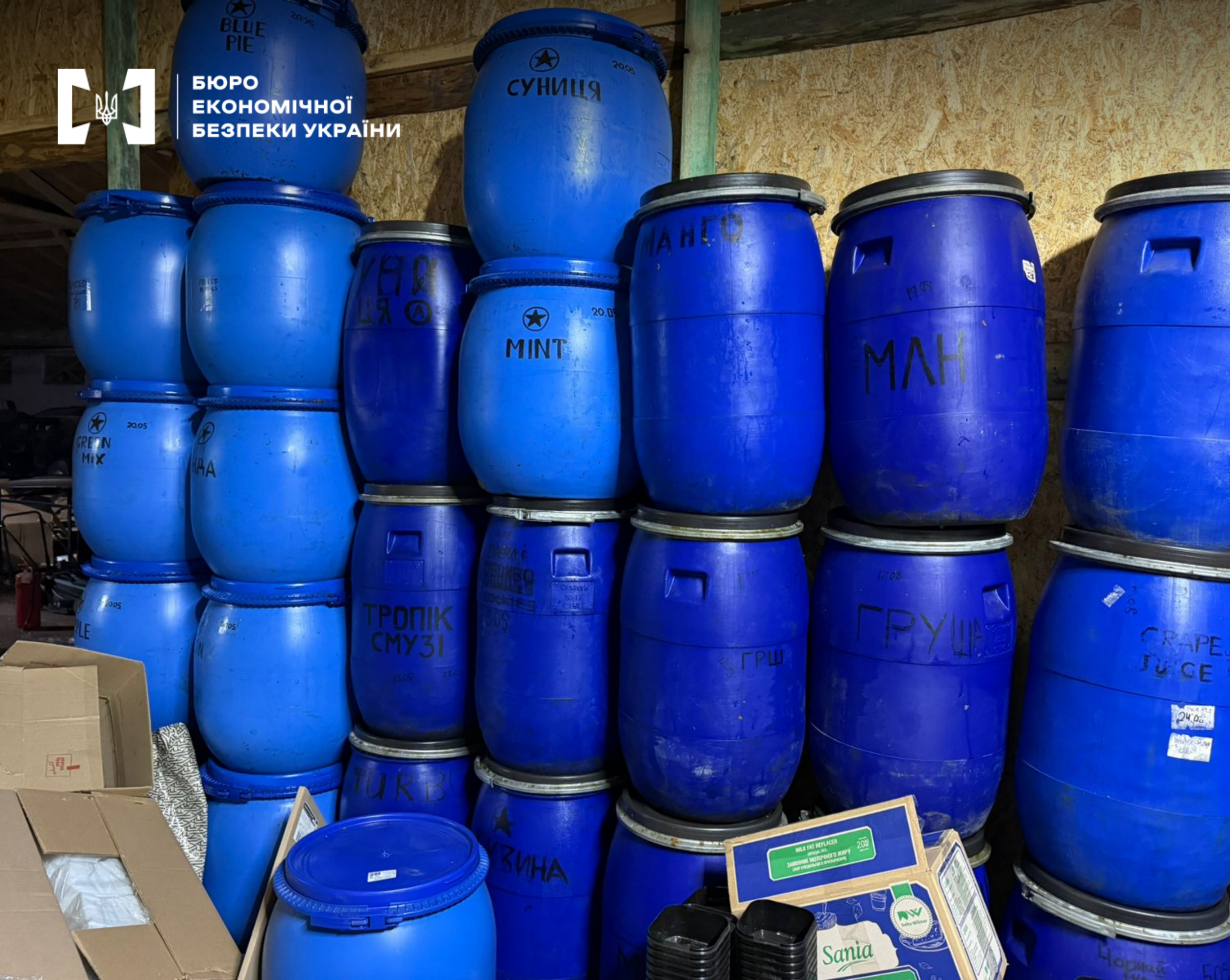 A warehouse with dozens of large blue plastic barrels, marked with hookah tobacco flavors in marker: 
