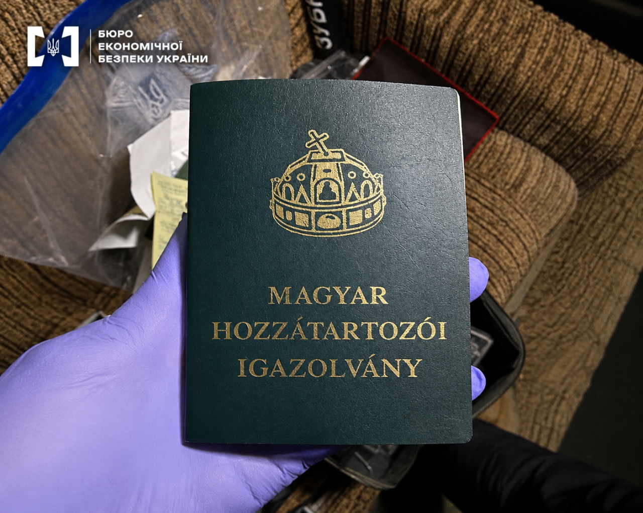 A hand in a blue glove holds a foreign identity document with a gold-embossed coat of arms.