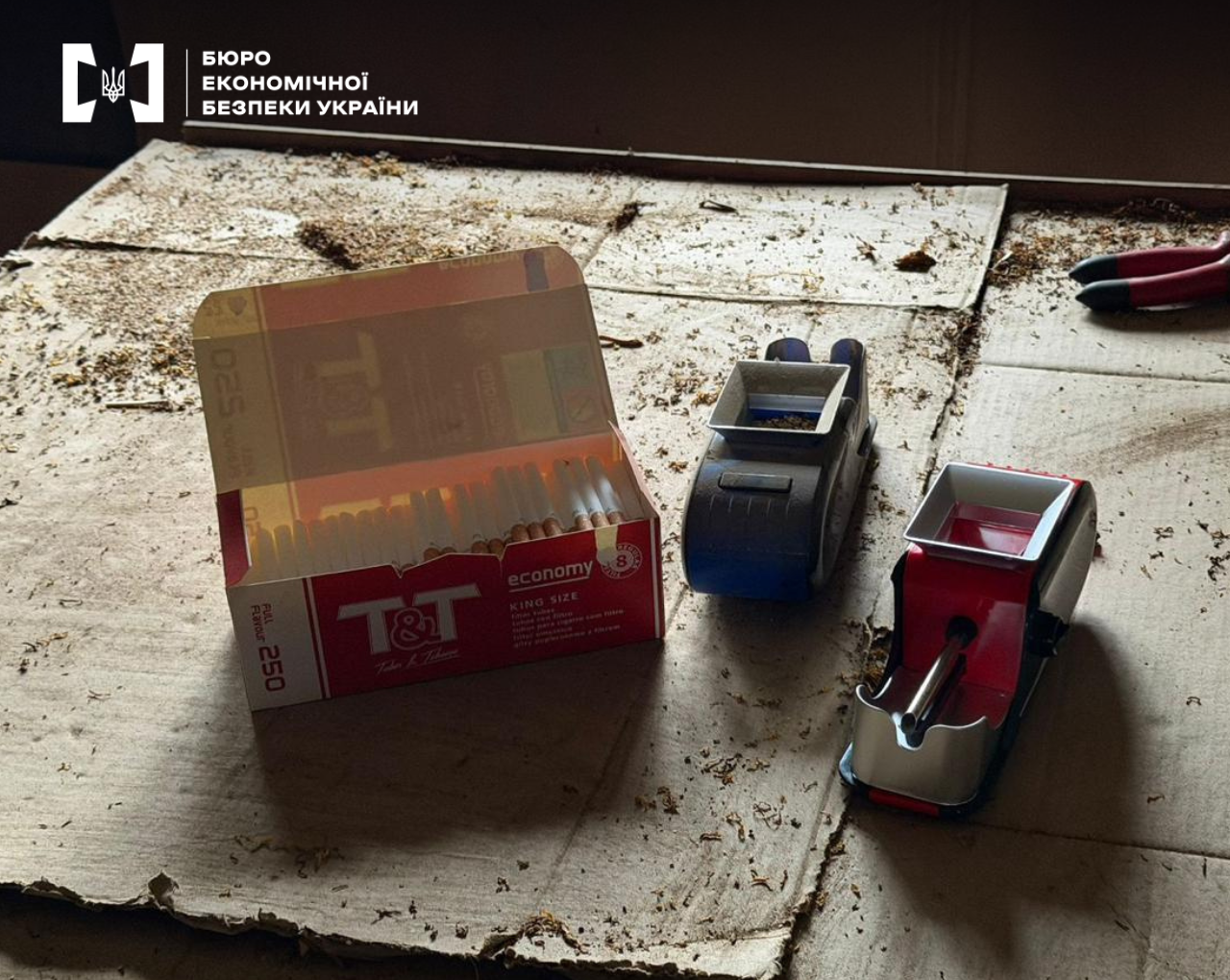 A box of 'T&T Economy' cigarettes on a piece of tobacco-covered cardboard. Next to it are two manual cigarette rolling machines.