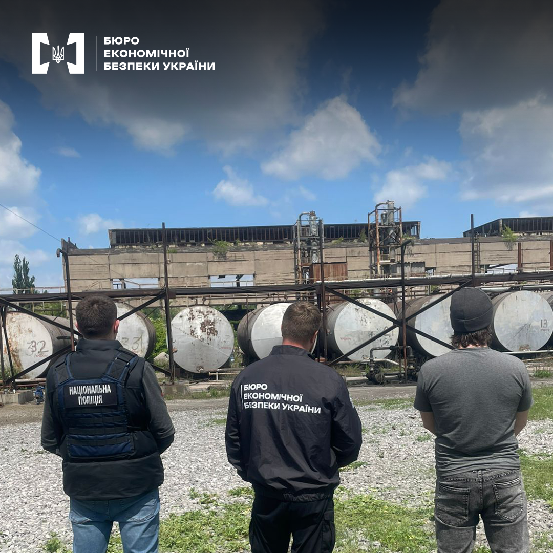 Працівники БЕБ та поліції на території нафтопереробного заводу. Промислова будівля, цистерни для зберігання палива. Блакитне небо з білими хмарами.