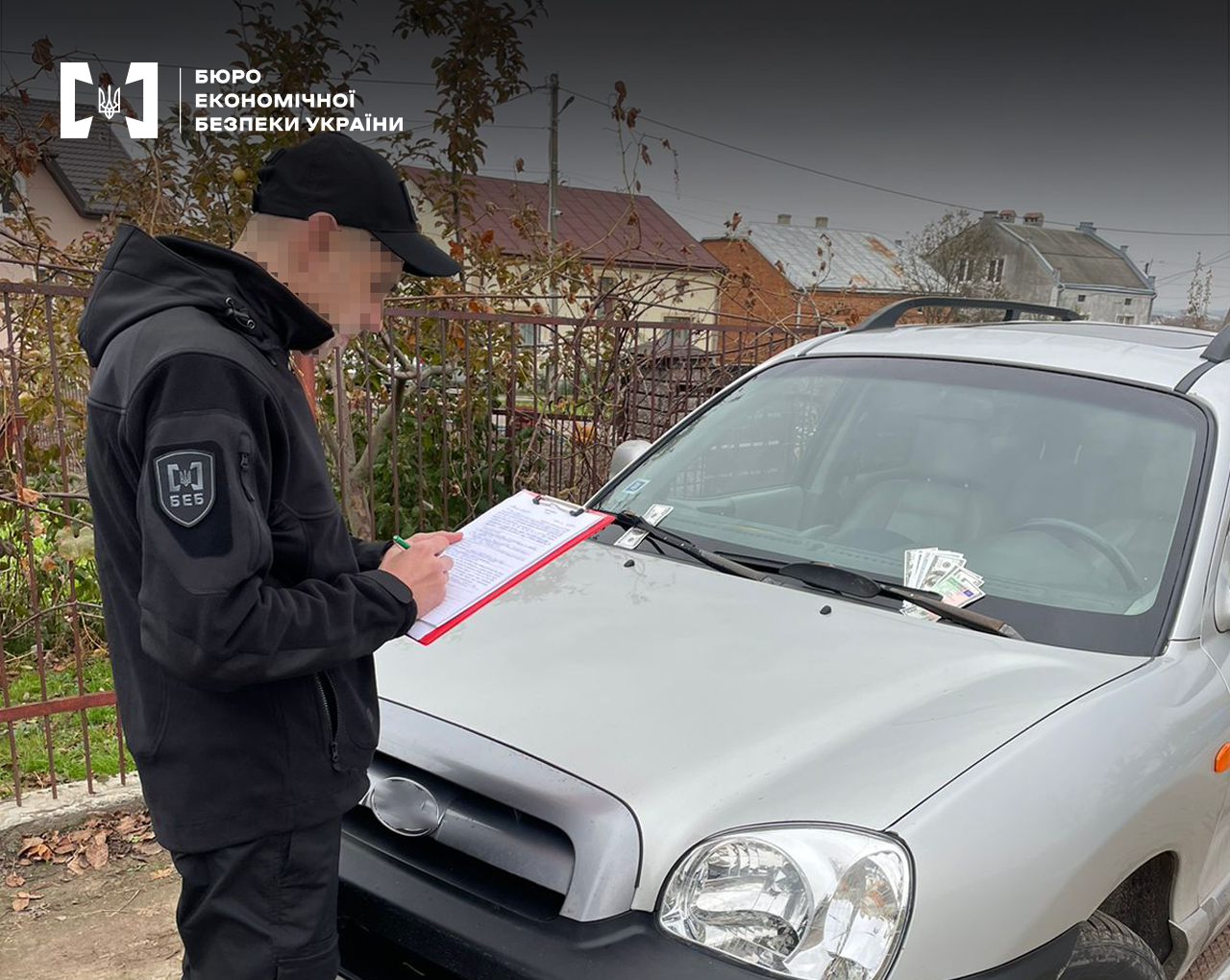 Приватний сектор. Детектив БЕБ поруч із легковим автомобілем оформляє документи. Під склоочисником авто долари та євро.