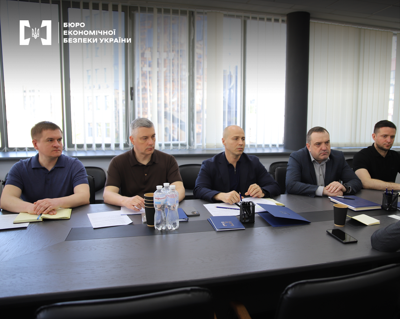 Учасники зустріч за столом. На столі вода в пляшках, стакани, записники, документи