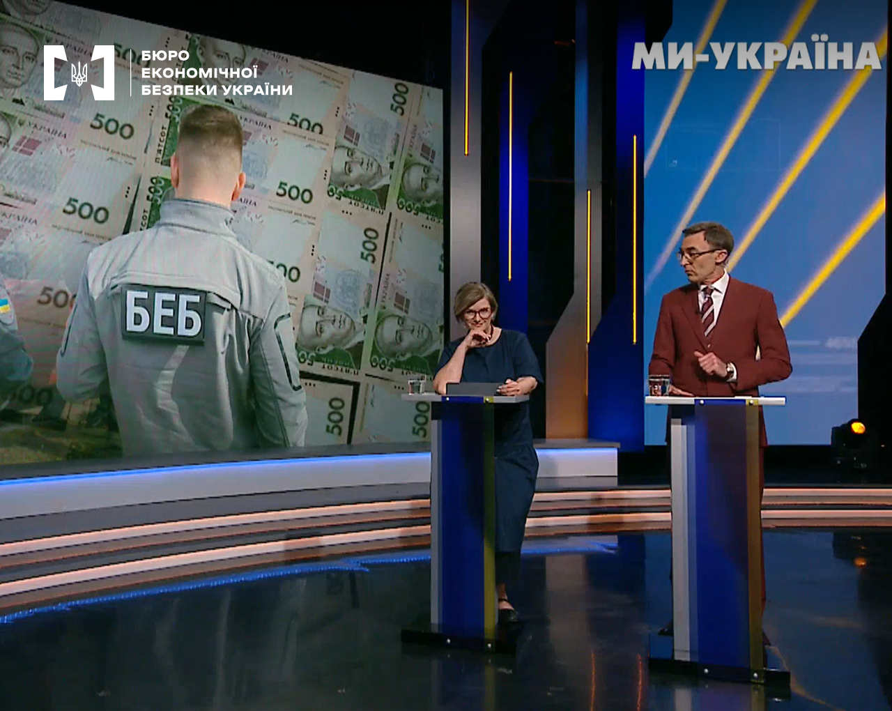 Студія телеканалу "МИ-Україна" в синіх кольорах: ведучі за стійками обговорюють діяльність БЕБ на фоні екрану з грошима.