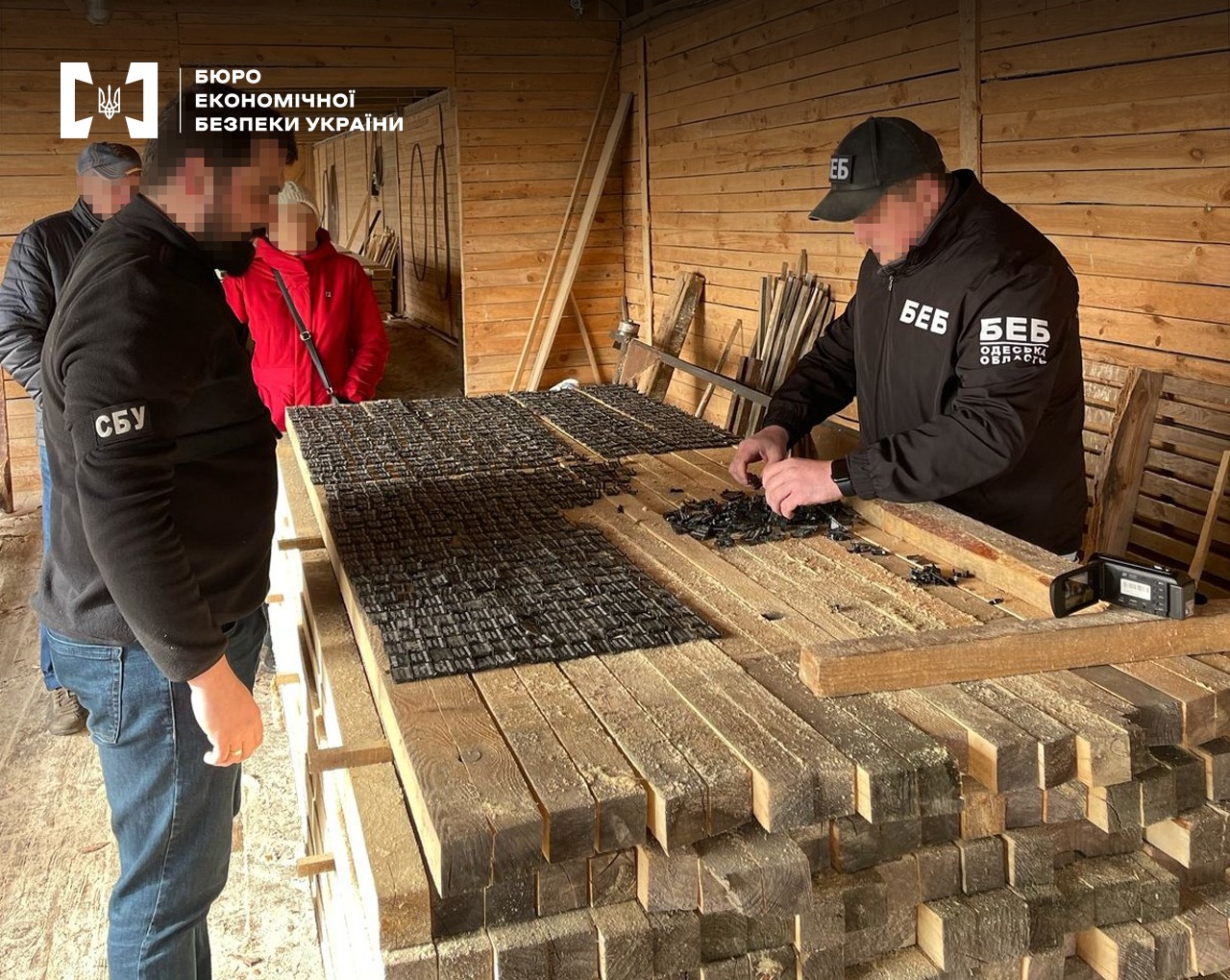 Працівники БЕБ та СБУ проводять слідчі дії на складі пиломатеріалів. Купа брусу, на якому чорні бірки для маркування деревини. Поруч чоловік та жінка.