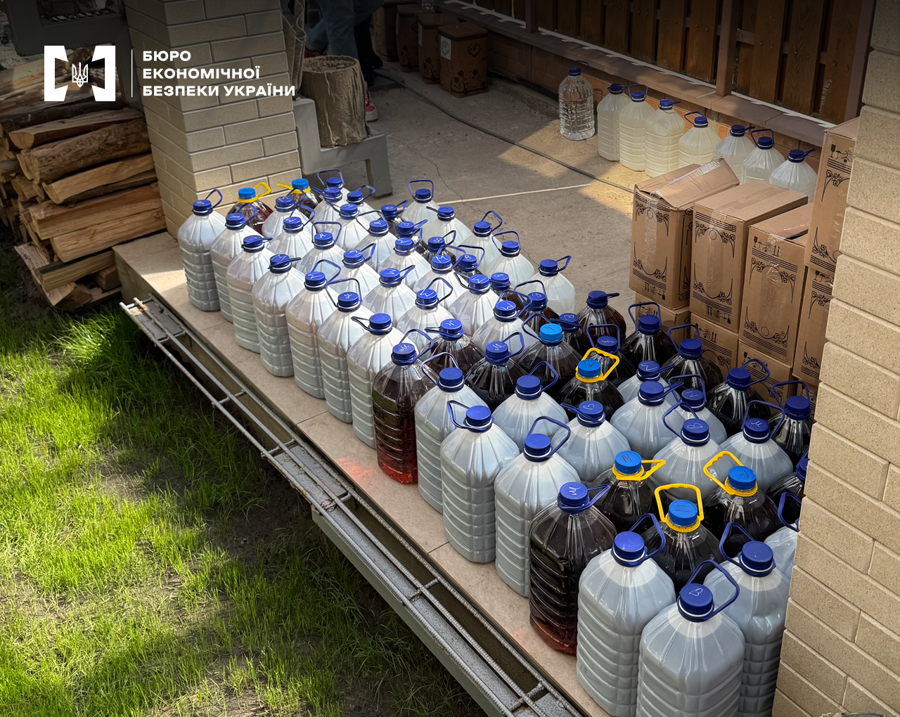Приватна прибудинкова територія, зелений газон. Шестилітрові ПЕТ пляшки з сурогатним алкоголем розставлені рядами. Поруч дрова, картонні коробки.