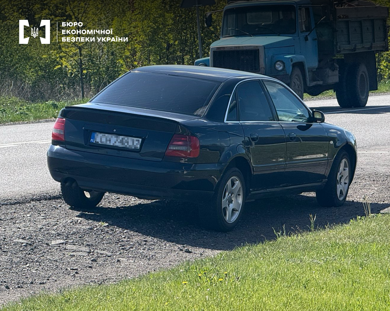 Темний легковий автомобіль на узбіччі дороги, повз проїжджає вантажівка.  З протилежної сторони дороги дерева. На узбіччі зелена трава.