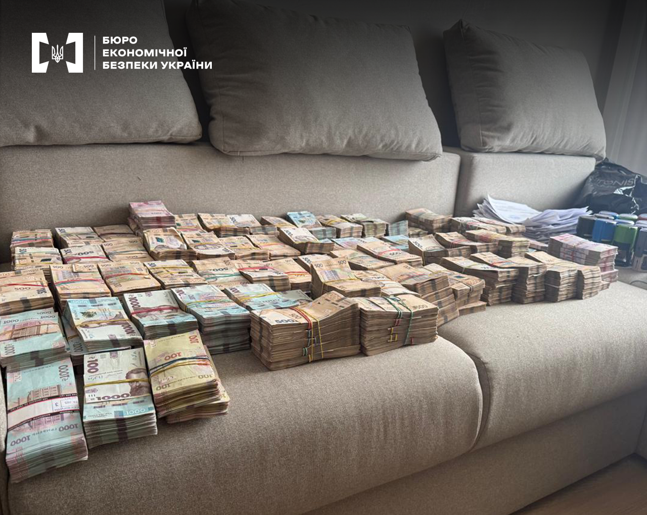 Величезні пачки гривневих купюр різного номіналу, документи, печатки, чорний пакет розкладені на  бежевому дивані.