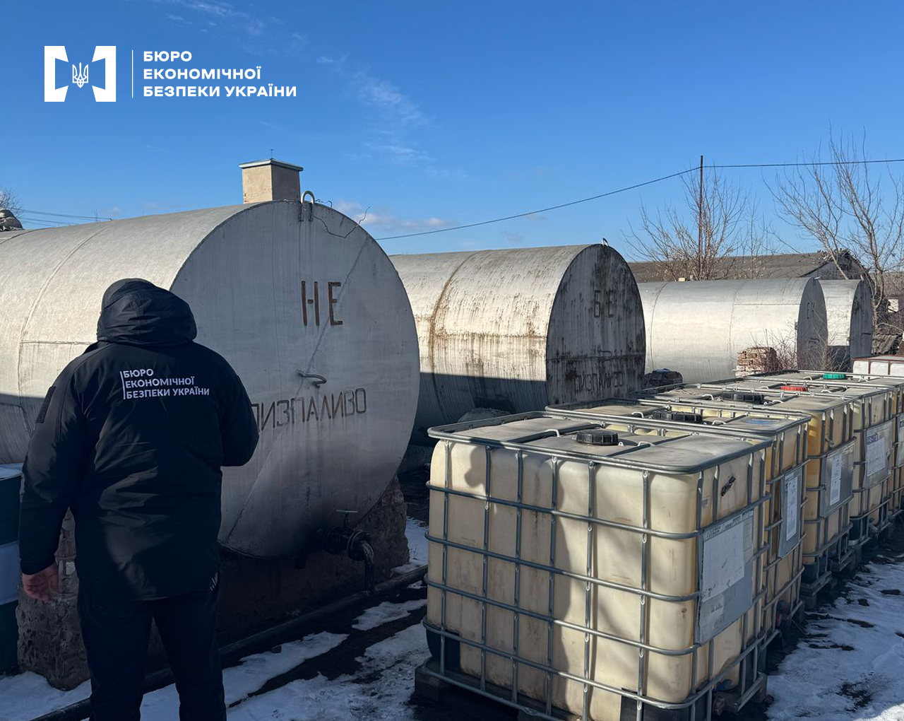 Детектив БЕБ оглядає металеві циліндричні резервуари з дизпаливом, що стоять один за одним. Поруч розміщені резервуари типу «єврокуб» для міні АЗС