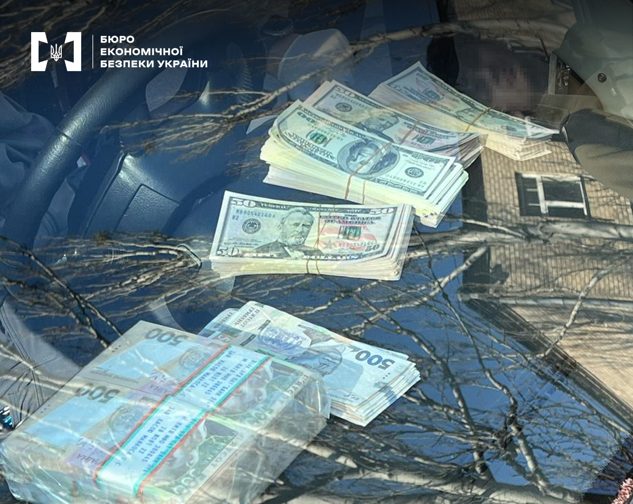 Пачки грошей номіналом 100 та 50 доларів США, 500 гривень на панелі приладів автомобіля. Фото через лобове скло, у відображенні гілки дерев, будівля.