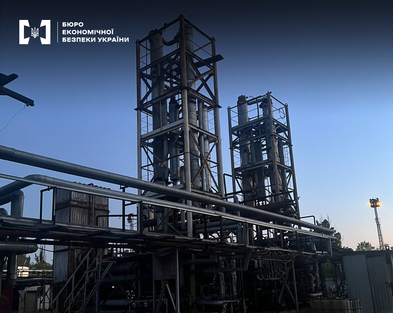 Великі металеві конструкції нафтопереробного заводу на тлі блакитного вечірнього неба.