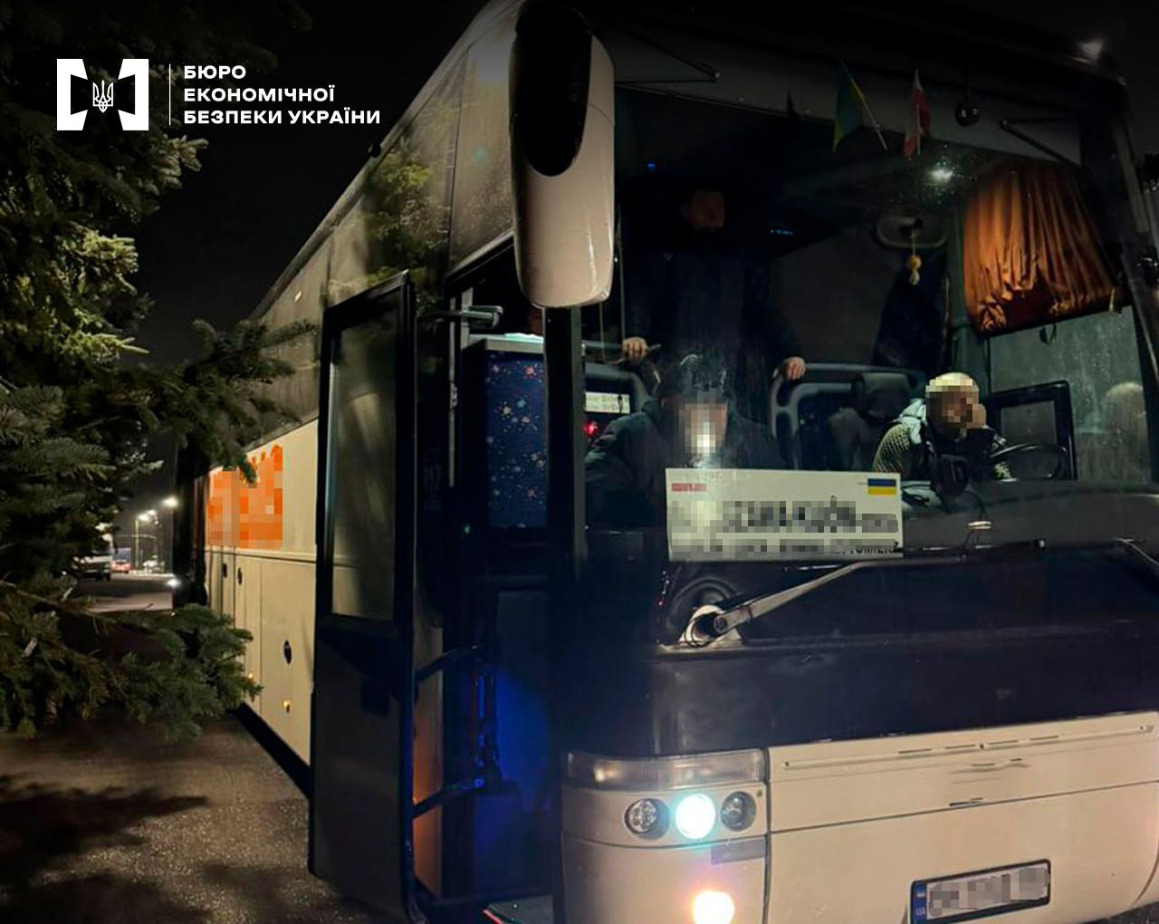 Пасажирський автобус далеких перевезень, у салоні водій та пасажири. Номерний знак та напрямок руху заблюрено. Темна пора доби.